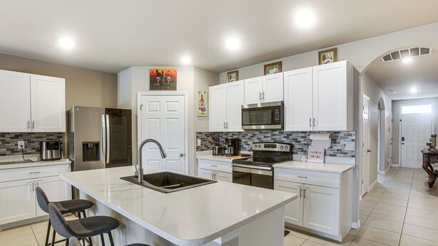 A modern kitchen with white cabinets, a stainless steel refrigerator, a microwave, and a large island with bar stools. - Lanai & Fenced Yard! Home 8 Mi to Charlotte Harbor (Punta Gorda)