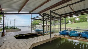 Ein überdachter Steg mit Booten, einem Holzdeck und Blick auf ein Haus und Bäume über dem Wasser.