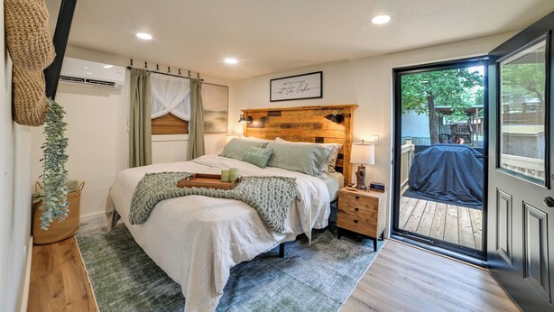 A bedroom with a bed, bedside table, lamp, and a view of the outdoors through a sliding door. - Hot Tub, Deck! Tiny Home Retreat By Kentucky Lake (Springville)