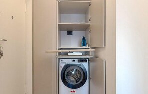 Ein kleiner Waschbereich mit einer Frontlader-Waschmaschine, einem kleinen Regal darüber und einem Schrank mit einem einzelnen Gegenstand auf der obersten Ablage.