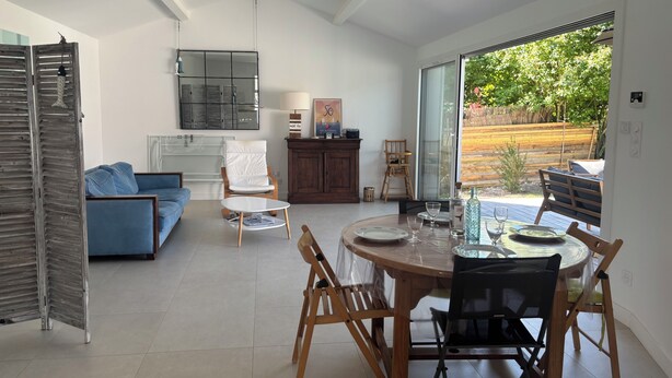 A modern living room with a blue sofa, a white armchair, a dining table with chairs, and a view of the outdoors through large windows. - Escapade rochelaise: new, modern house (La Rochelle)