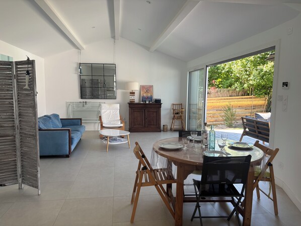 Ein modernes Wohnzimmer mit einem blauen Sofa, einem weißen Sessel, einem Esstisch mit Stühlen und Blick ins Freie durch große Fenster.