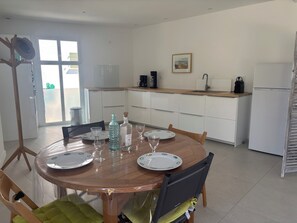 Ein Essbereich mit einem runden Tisch, gedeckt für vier Personen. Im Hintergrund ist eine Küche erkennbar, und eine Glastür öffnet sich zu einem Außenbereich.