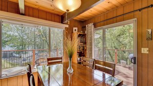 A wooden-paneled room with a dining table, chairs, a vase with plants, and large windows offering a view of trees.