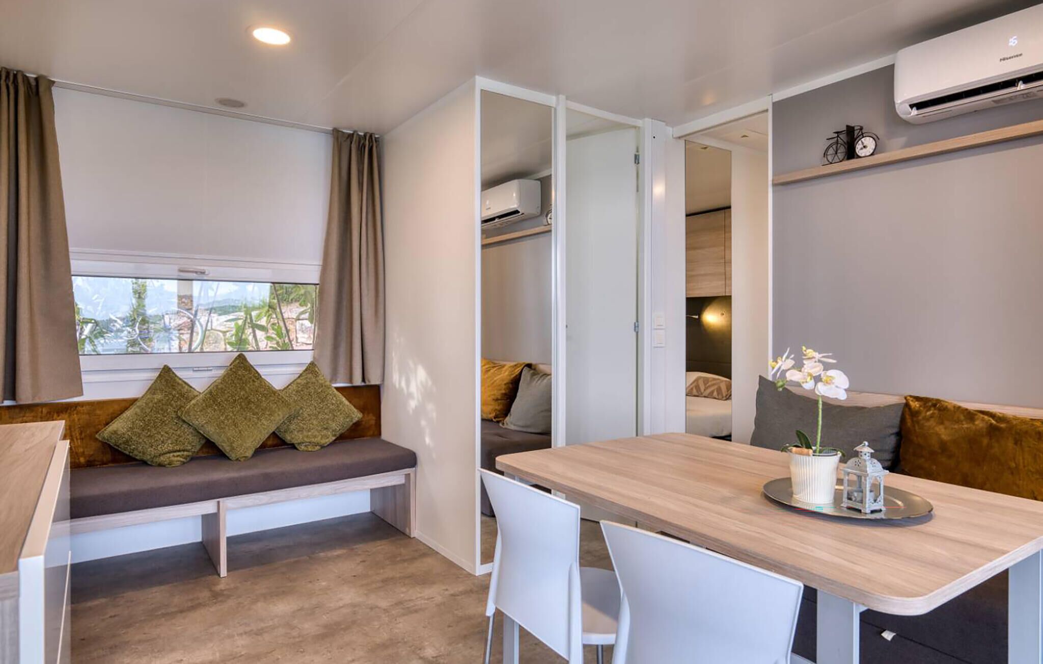 Habitación compacta con un banco, una mesa, sillas, una orquídea en maceta y un aire acondicionado de pared.