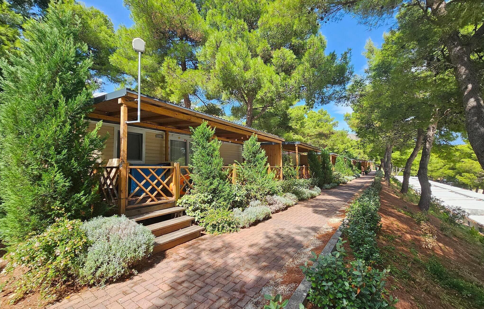 Une rangée de cabanes en bois, reliées par un chemin pavé de briques, et entourées d’arbres.