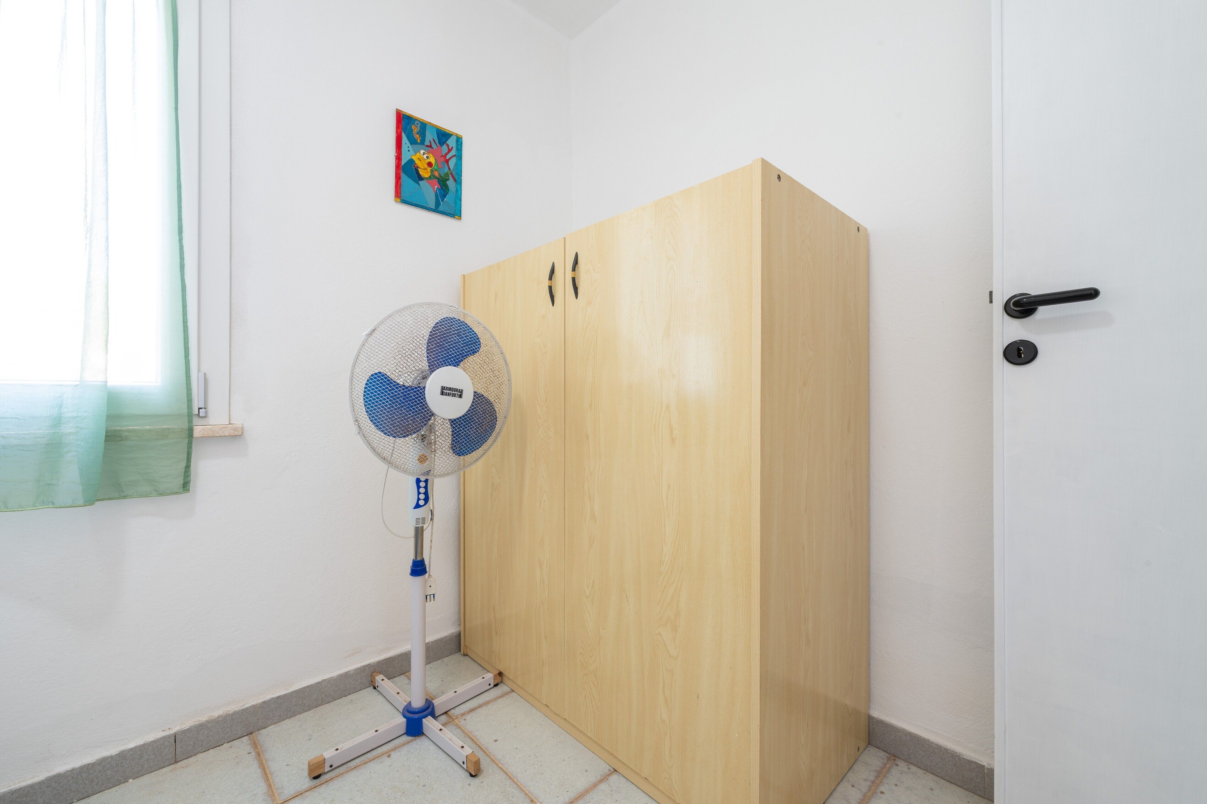 Una habitación con un armario de madera, un ventilador y una ventana con cortina verde.