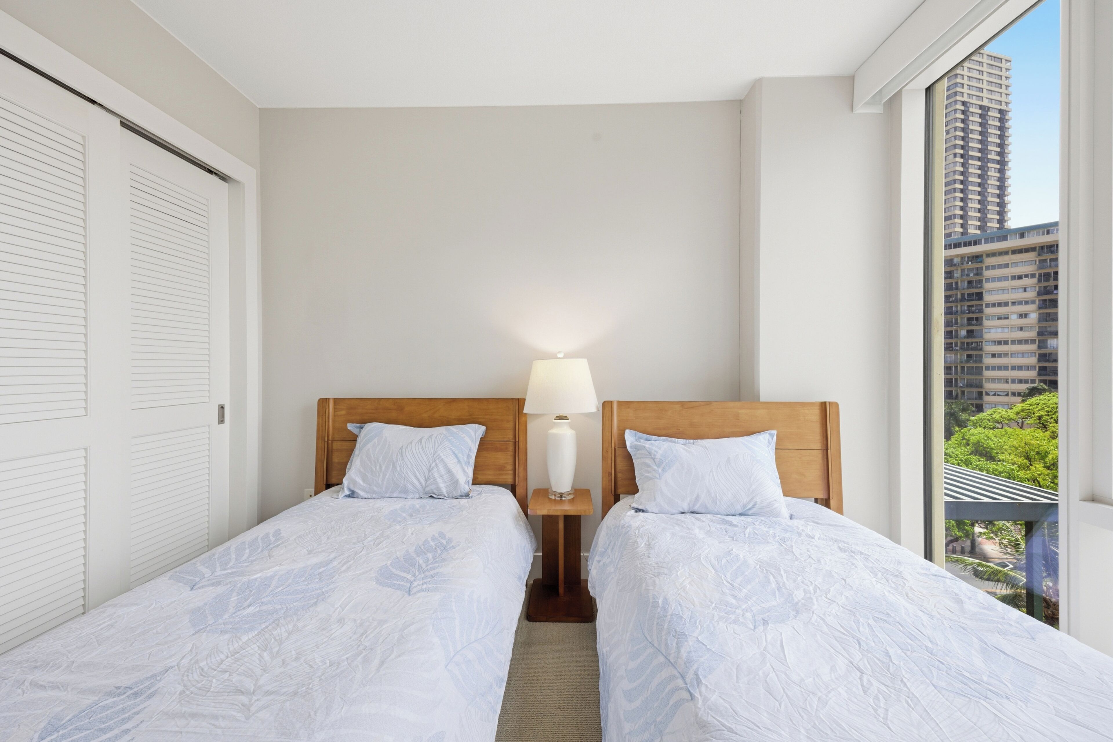A hotel room with two single beds, a nightstand with a lamp, a large window with a city view, and a sliding door closet.