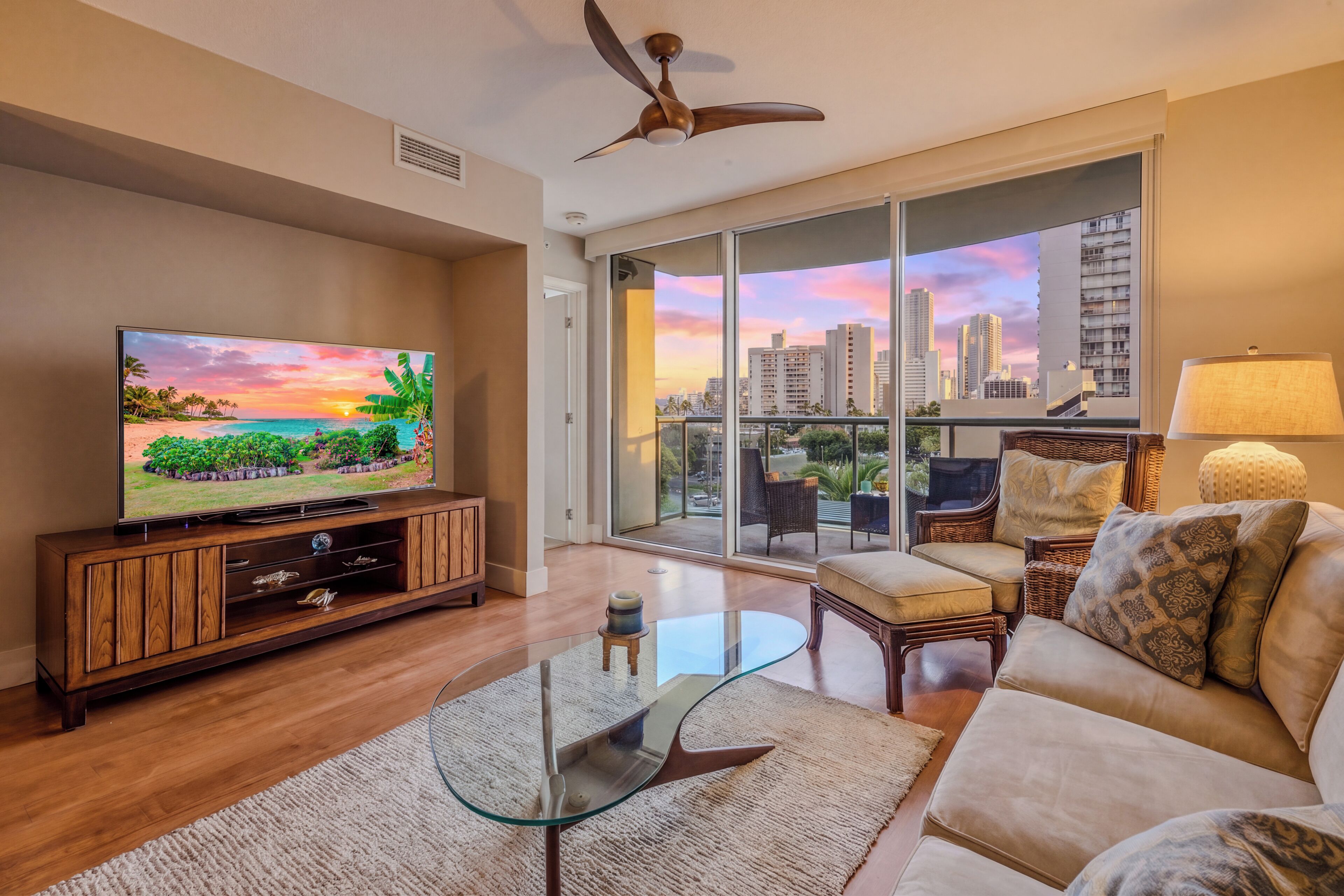 A living room with a TV, glass coffee table, sofa, and a view of the cityscape through large windows.