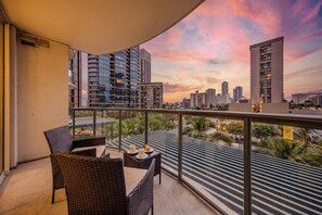Ein Balkon mit Korbmöbeln, einem Tisch mit Kerze und Blick auf die Stadt bei Sonnenuntergang.
