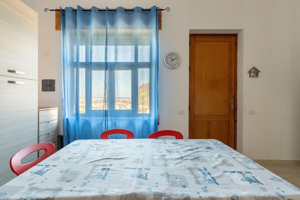A room with a table, chairs, a wooden door, a window with blue curtains, and a wall clock.