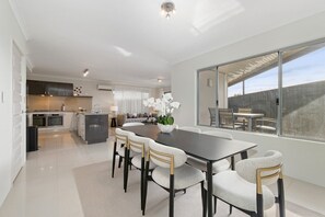 A modern kitchen with a central island, a dining area with a large table and chairs, and a view of the outdoors through large windows. - The Grand Aveley House (Aveley)