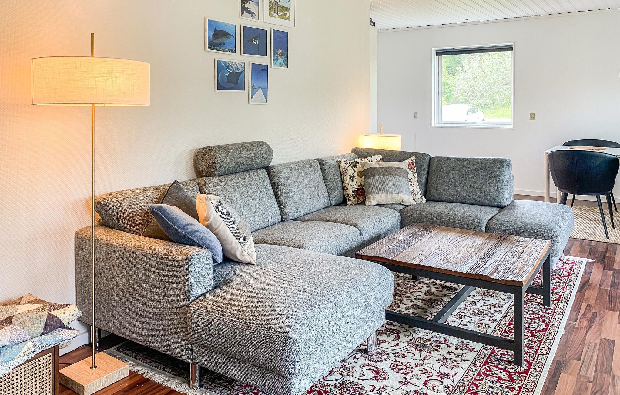 Ein Wohnzimmer mit Ecksofa, Couchtisch, Stehlampe, Teppich mit Muster und einem Fenster mit Blick auf Grünflächen.