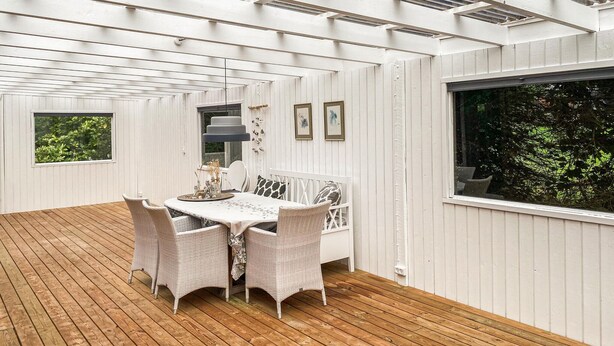 Une pièce avec une terrasse en bois, un lit blanc, des meubles en osier et des tableaux encadrés accrochés au mur.