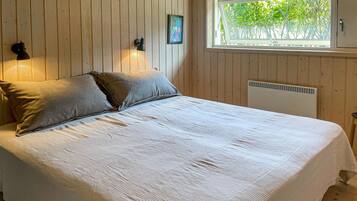Un lit recouvert d’une couverture blanche, deux oreillers, une lampe fixée au mur, un tableau encadré et une fenêtre donnant sur des arbres.