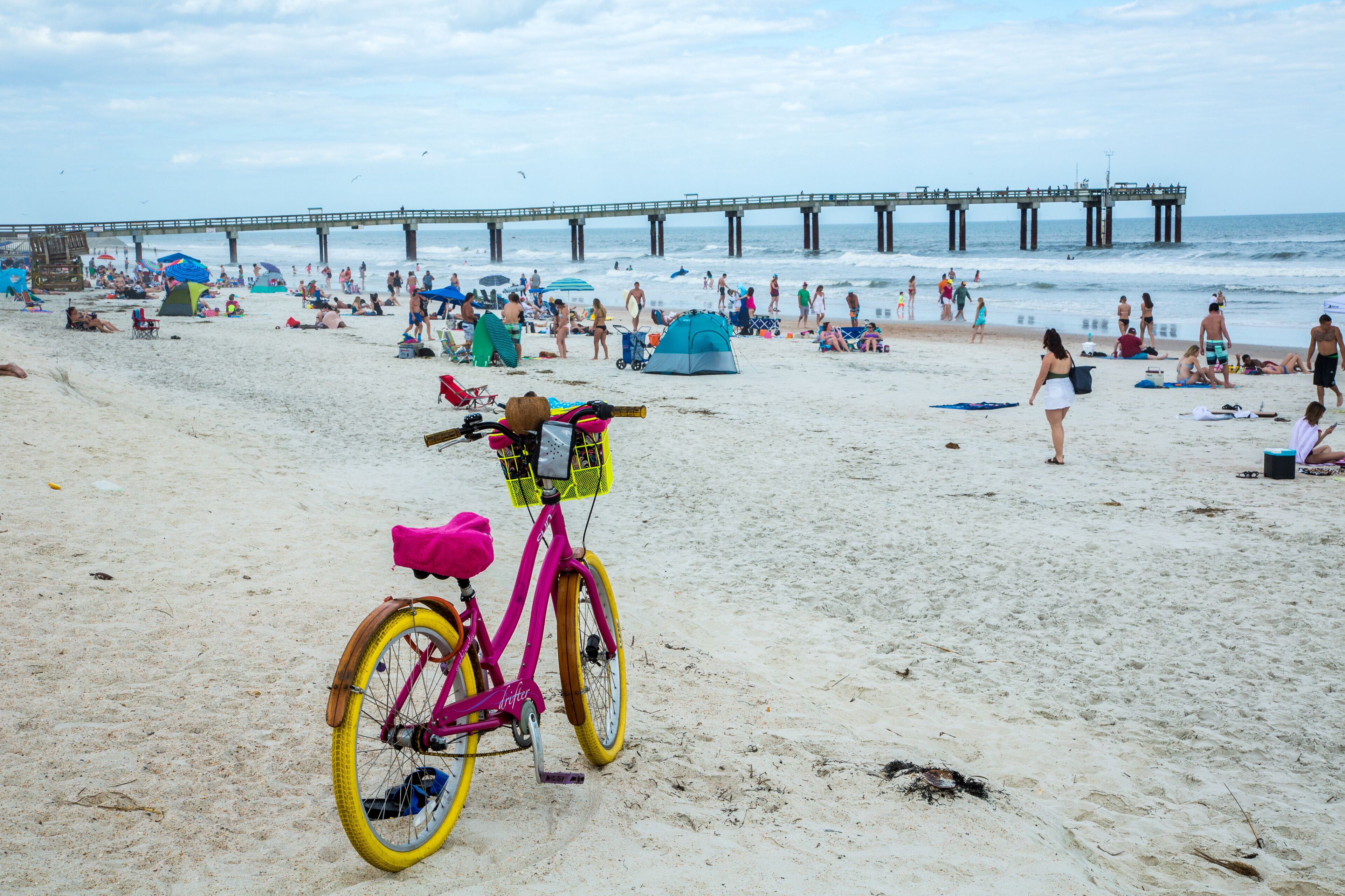 A beach with a pier in the background, people relaxing, and a pink bicycle with a basket in the foreground.
