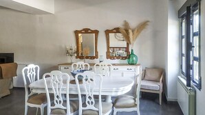 Een eetgedeelte met een witte tafel en stoelen, twee spiegels, een vaas met gedroogde planten en een raam met zonwering.