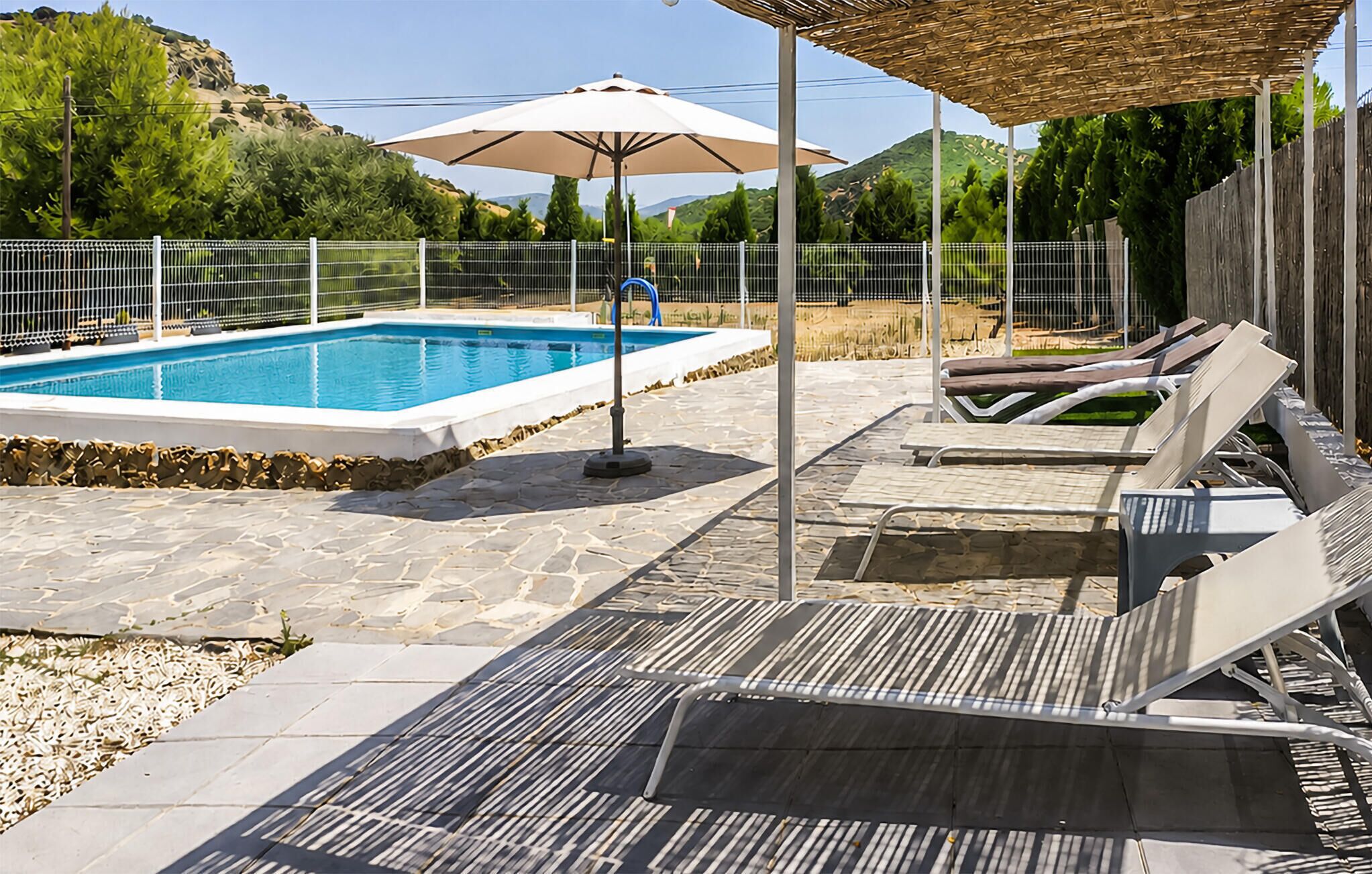 Una piscina con lettini e ombrelloni, circondata da un solarium in pietra e una recinzione.