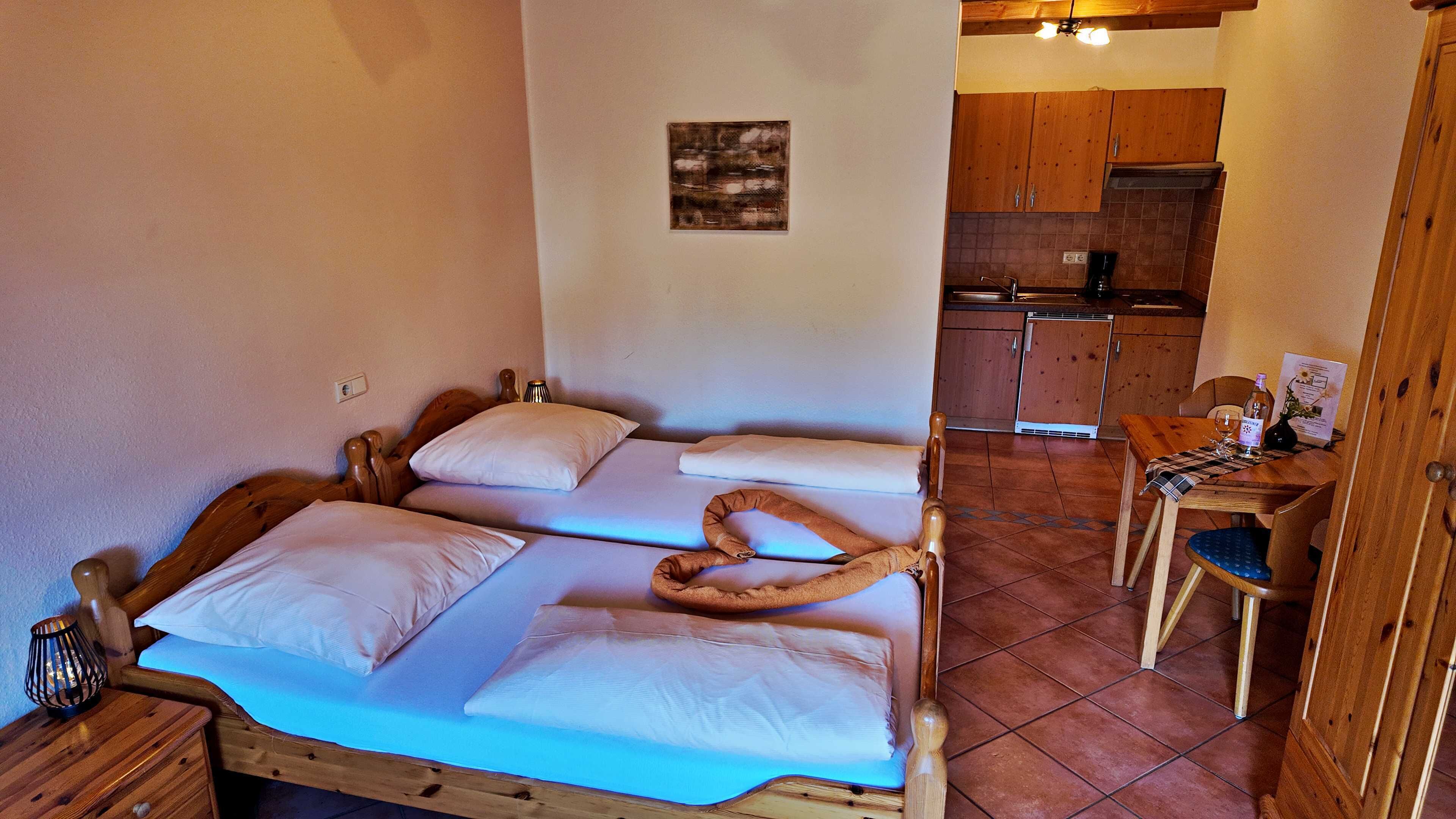 A cozy room with two wooden beds, a small table with a chair, and a kitchen area with wooden cabinets.