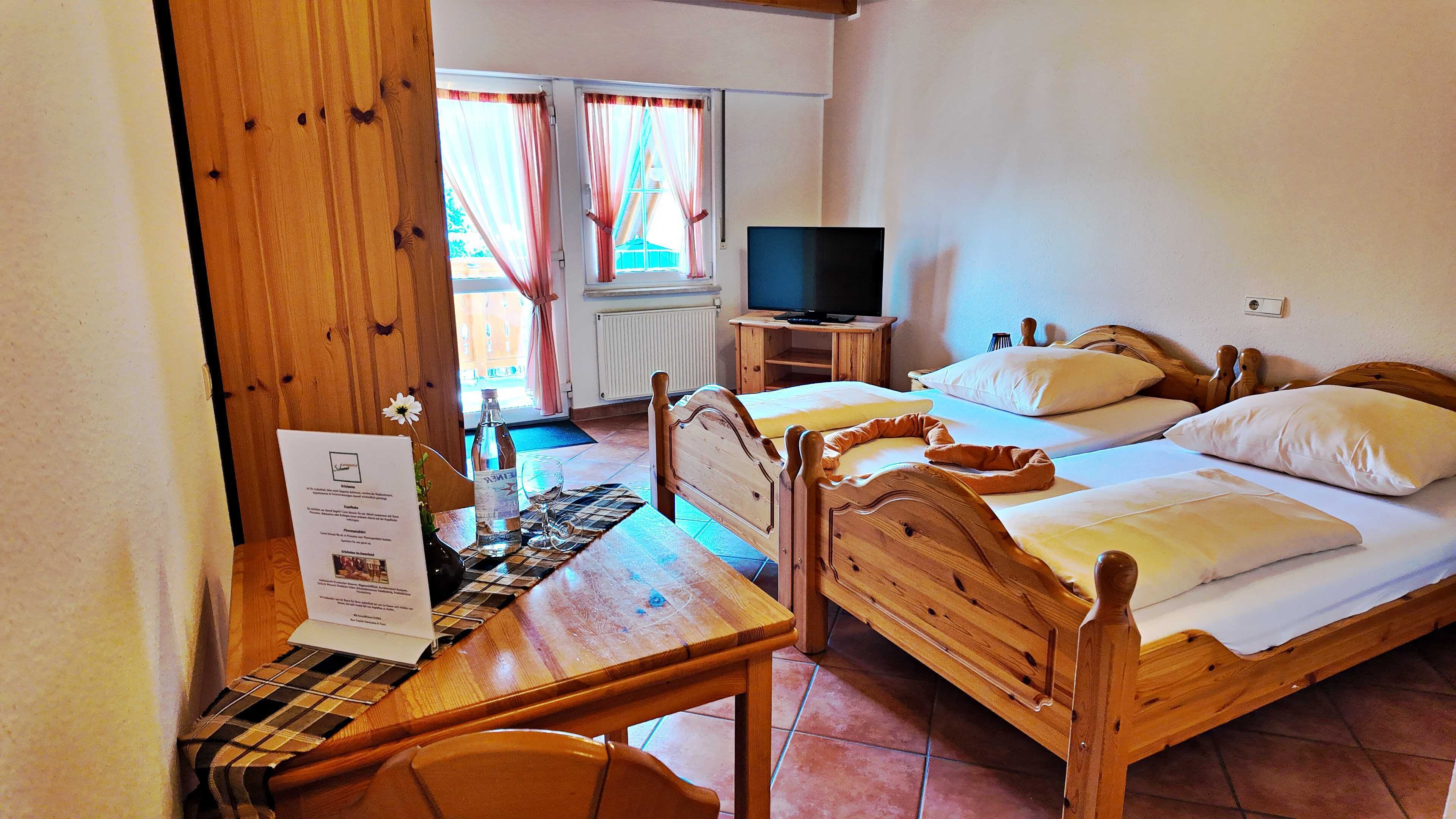 Una habitación con dos camas de madera, un televisor sobre un mueble de madera, una mesa con mantel individual y una ventana con cortinas.