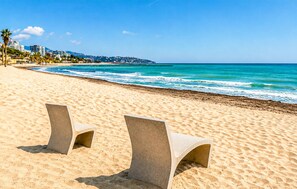 Ein Strand mit zwei Betonstühlen, wolkenlosem, blauem Himmel und einer entfernten Skyline.