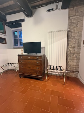 Ein Zimmer mit einem Holzkasten, einem Fernseher darauf, einem Heizkörper und einem Fenster mit Blick auf Bäume.