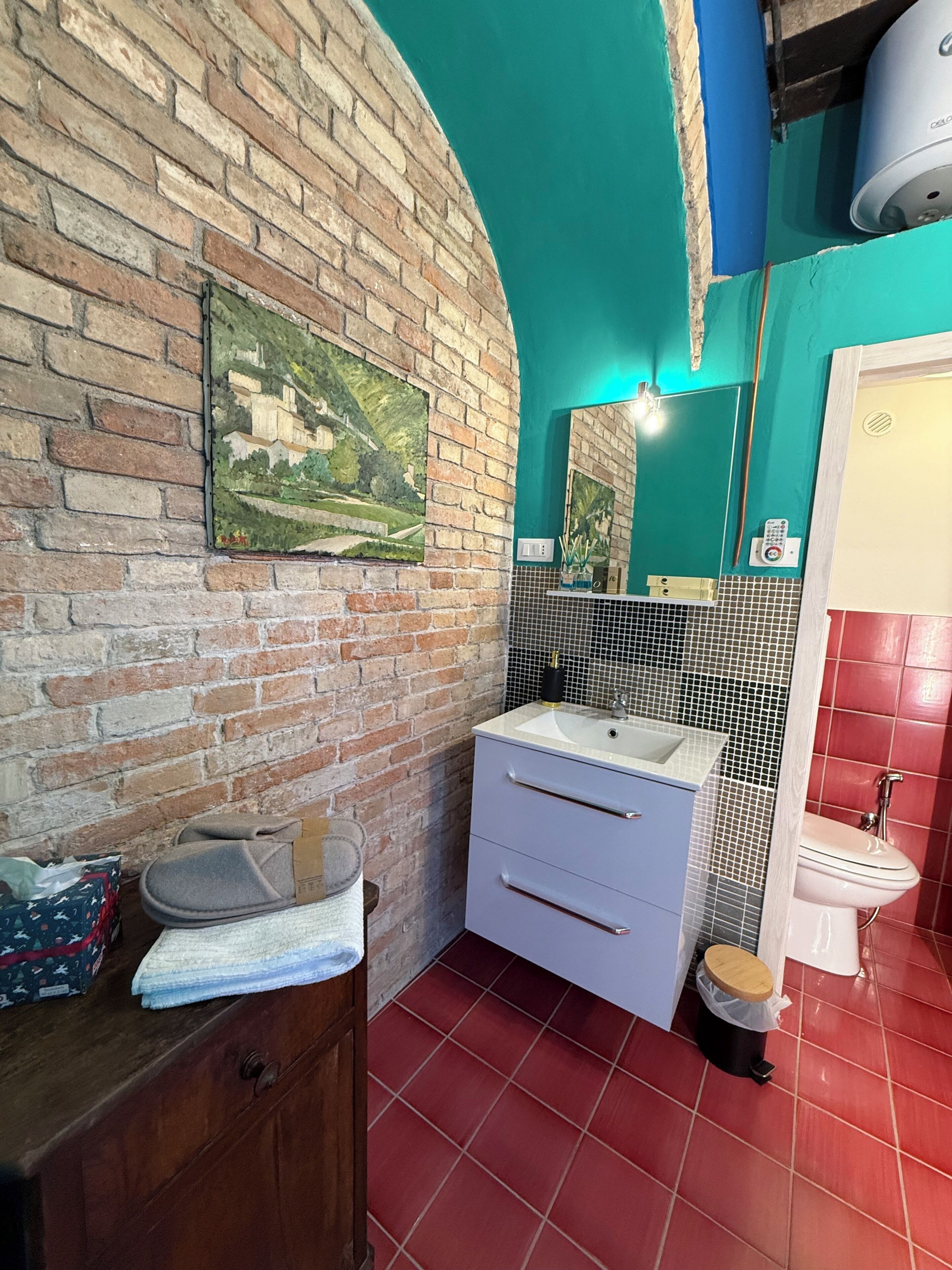 Un baño con pared de ladrillo, un lavabo, un espejo, un inodoro y un pequeño cubo de basura.