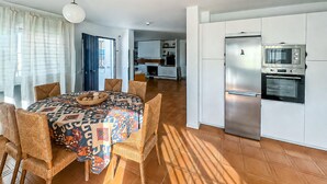 Una cocina moderna con horno empotrado y refrigerador, un comedor con mantel estampado y sillas de mimbre, y vista al área de estar.