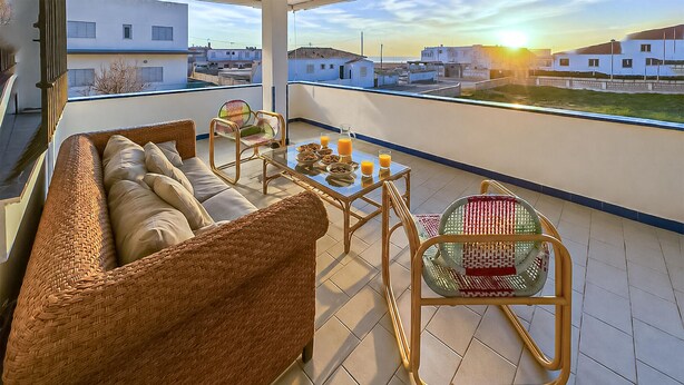 Un balcón con mobiliario de mimbre, una mesa de vidrio y vistas a edificios al fondo.