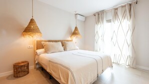 Ein Schlafzimmer mit einem Bett, zwei Pendelleuchten, einem Fenster mit Vorhängen und einem kleinen Korbwäscher.