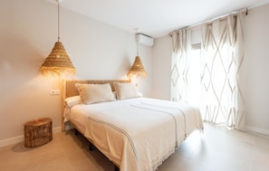 Ein Schlafzimmer mit einem Bett, zwei Pendelleuchten, einem Fenster mit Vorhängen und einem kleinen Korbwäscher.