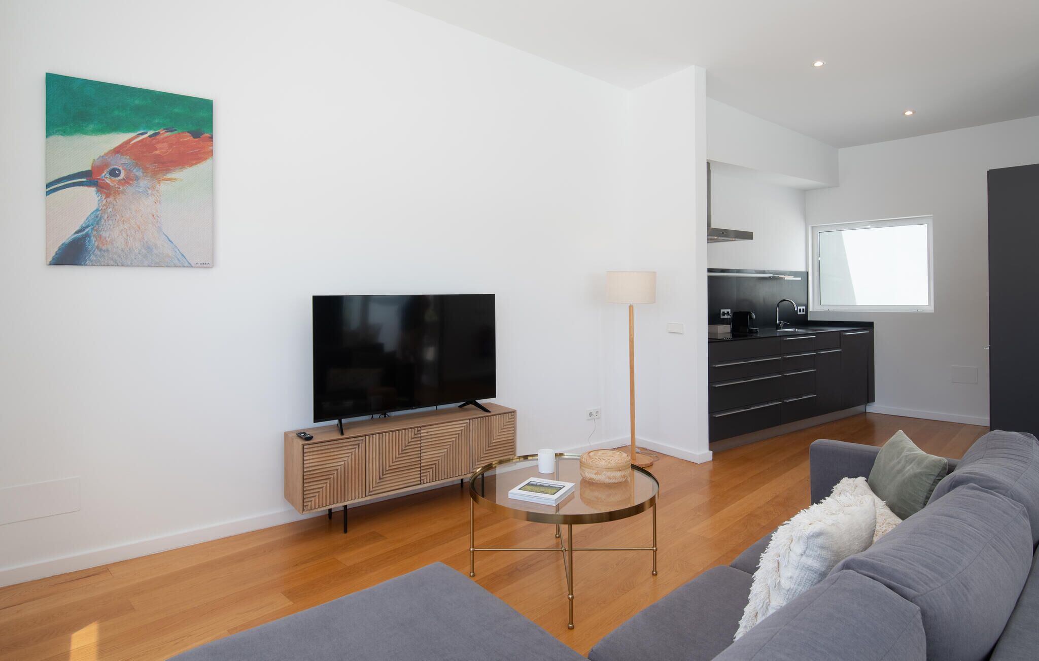 A modern living room with a flat-screen TV, a wooden cabinet, a round glass coffee table, a grey sofa, and a kitchen area with a window.