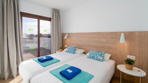 Ein Hotelzimmer mit zwei Betten, einem Balkon, einem Holzkopfende, einem kleinen Nachttisch und einer Wandlampe.