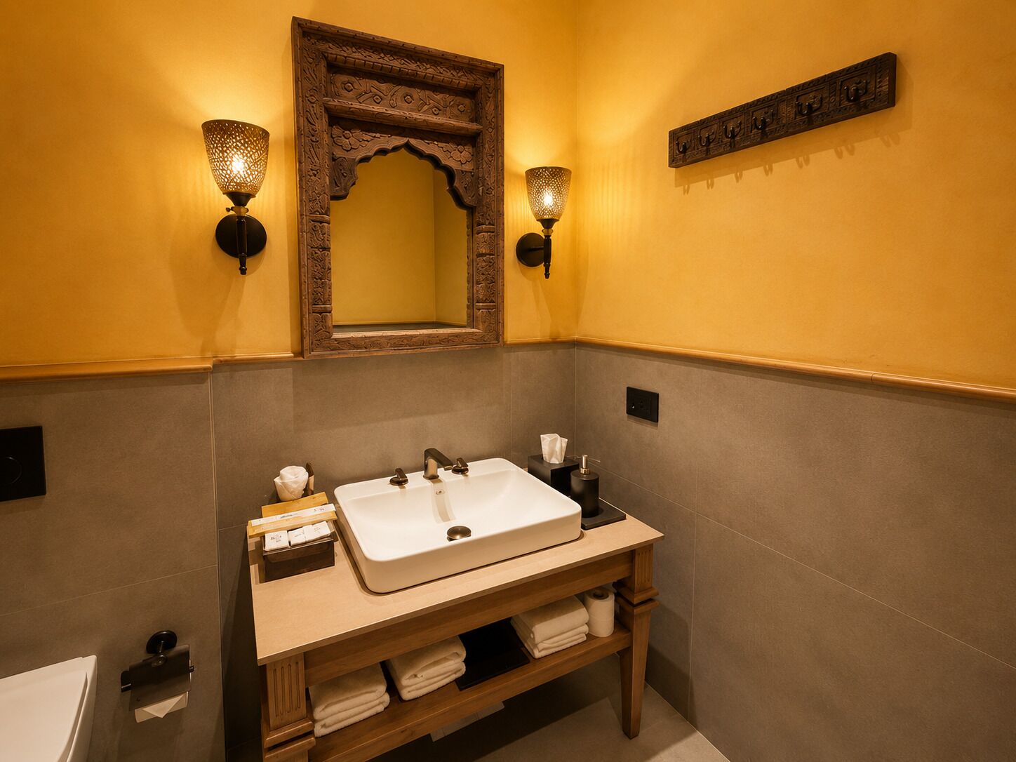 Un baño con un lavabo blanco, dos luces empotradas en la pared, un mueble de tocador de madera y un espejo con detalles ornamentales.