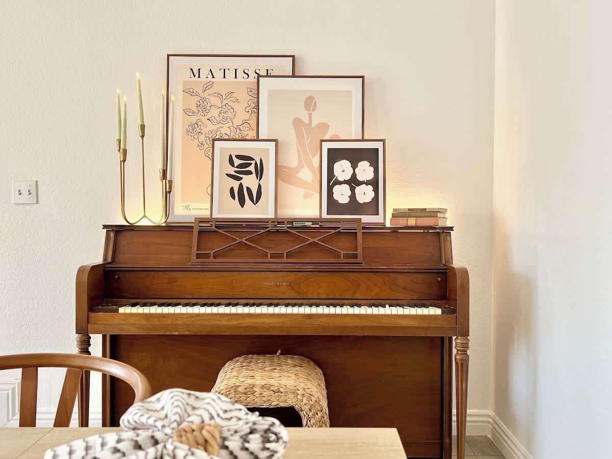 A piano with framed artwork above it, a woven basket on the piano, and a chair in front.