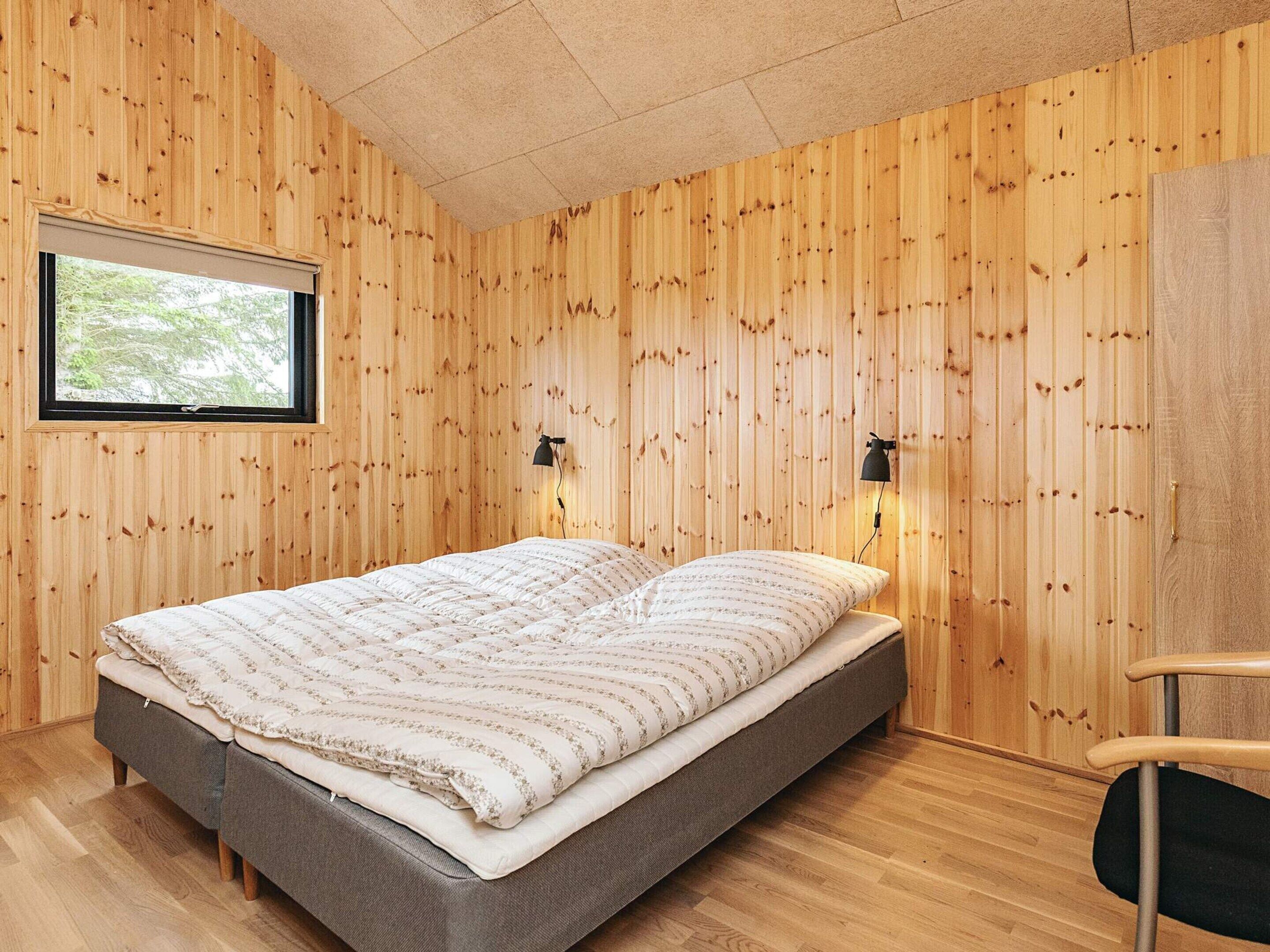 A room with wooden panel walls, a bed with a patterned bedspread, two wall-mounted lamps, and a window with a view of trees.