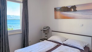 Ett hotellrum med en säng, en målning som föreställer ett kustlandskap, ett fönster med gardiner och utsikt över havet.