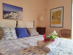 Un letto rifatto con cura, copriletto fantasia, un cuscino, un comodino con una lampada e un quadro incorniciato al muro.