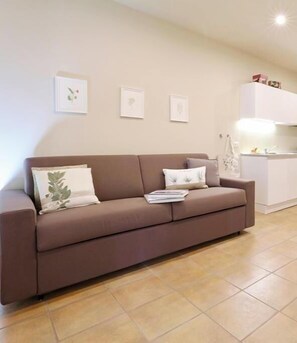 Ein braunes Sofa mit Kissen und einer Zeitschrift, ein weißes Schränkchen mit Gegenständen darauf und ein gefliester Boden.