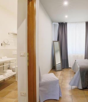 Ein Hotelzimmer mit Holztür, Waschbecken in Weiß, einem Spiegel, zwei Betten mit grauen Bettwäsche und einem Fenster mit Vorhängen.