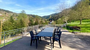 An outdoor dining area with a table and chairs, surrounded by a metal railing, overlooking a valley with houses and trees. - Les Privilèges family home in the heart of nature, 5 minutes from the town center (Gérardmer)