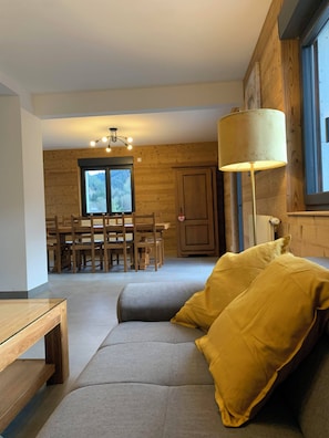 Ein modernes Wohnzimmer mit einem grauen Sofa, gelben Kissen, einem Holzesstisch und Stühlen. Ein großes Fenster bietet einen Blick ins Freie.