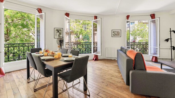 Een eetgedeelte met een tafel gedekt voor twee personen, een bank en een balkon met uitzicht op bomen.