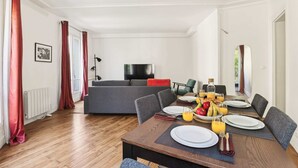 Een eetgedeelte met een houten tafel gedekt voor het ontbijt, een grijze bank op de achtergrond en een televisie aan de muur.
