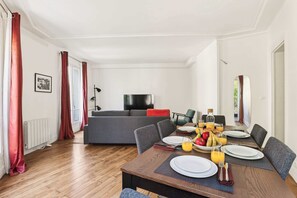 Ein Essbereich mit einem Holztisch, der für ein Frühstück gedeckt ist, einer grauen Couch im Hintergrund und einem Fernseher an der Wand.