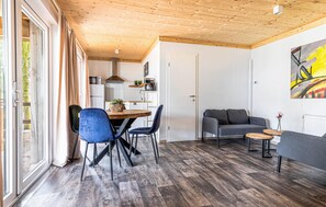 Ein modernes Wohnzimmer mit Holzboden, einem Essbereich mit Holztisch und blauen Stühlen, einem grauen Sofa und einem Bild an der Wand.