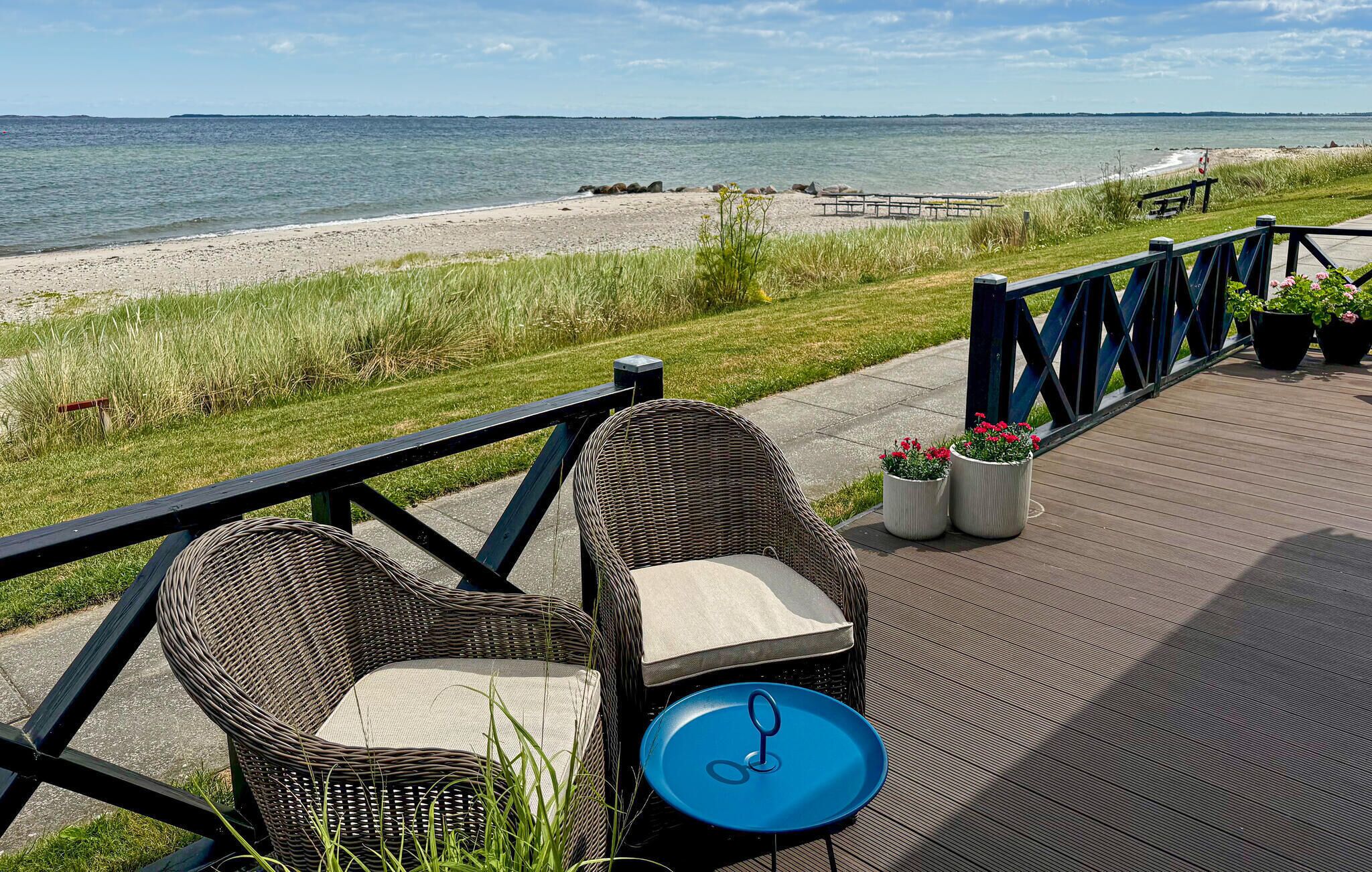 Une terrasse dotée de chaises en osier et d’une table bleue, offrant une vue sur une plage avec une promenade en bois et une rambarde.