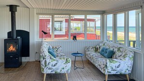 Una habitación acogedora con una estufa de leña, dos sofás con estampado floral, una mesita y grandes ventanas con vistas a la playa.