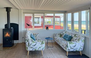 Ein gemütliches Zimmer mit Kaminofen, zwei Sofas mit Blumenmuster, einem kleinen Tisch und großen Fenstern mit Blick auf einen Strand.