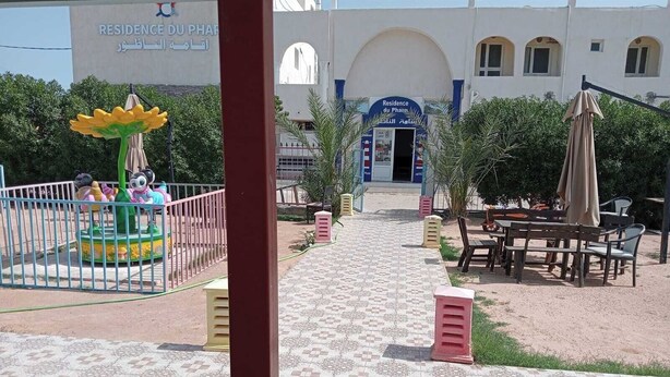 A courtyard with a children's play area, outdoor seating, and a building with a sign reading 'Residence du Phare'. - Résidence Le Phare (Djerba Midun)